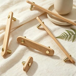 May include: A selection of wooden cabinet handles with brass fittings. The handles are rectangular with rounded ends and are made of light-coloured wood. The brass fittings are cylindrical. The handles are arranged on a white surface.