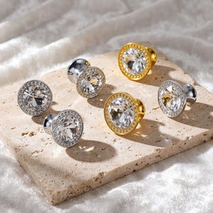 May include: A collection of six circular cabinet knobs featuring a central faceted clear crystal stone surrounded by a metallic rim. The set includes three silver-toned knobs and three gold-toned knobs, each with a textured border.