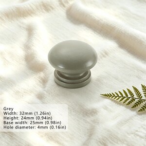Puede incluir: Un pomo de mueble gris, redondo y liso, con una base plana. El pomo tiene una anchura de 32 mm, una altura de 24 mm, una anchura de base de 25 mm y un di&aacute;metro de orificio de 4 mm, seg&uacute;n el texto impreso en la imagen.