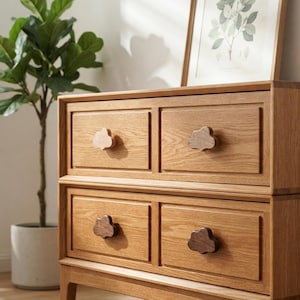May include: A light brown wooden dresser featuring four rectangular drawers with raised borders. Each drawer is equipped with a dark brown wooden knob shaped like a cloud. The dresser has a natural wood grain finish and stands on four short legs.