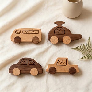 Puede incluir: Cuatro vehículos de juguete de madera: un autobús, un coche, un sedán y un camión. El autobús y el camión son de madera de color claro, mientras que el coche y el sedán son de madera oscura. Cada uno tiene ruedas de madera y detalles de ventanas grabados. Un helecho y un jarrón beige están en el fondo.