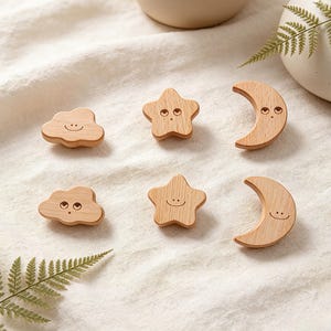 May include: Wooden cloud, star, and moon shapes with smiling faces. The shapes are made of light-colored wood and are arranged on a white fabric surface. The shapes are likely decorative items or toys.