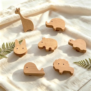 May include: A set of seven light-colored wooden cabinet knobs shaped like various animals, including a giraffe, elephant, rabbit, hippopotamus, turtle, whale, and lion. Each knob features a smooth, natural wood grain finish and a simple silhouette.