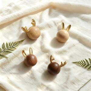 May include: Four spherical knobs featuring gold-toned metal antlers or ears. Two knobs are crafted from light-colored wood, while the other two are made from a darker, rich-toned wood. Each piece has a smooth, rounded base and metallic accents.