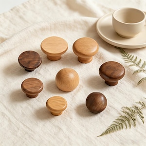 May include: A collection of wooden cabinet knobs in various shades of brown and natural wood tones. The knobs have a round or mushroom-shaped design, with some featuring a flat top and others a rounded shape. The knobs are arranged on a cream-colored fabric.