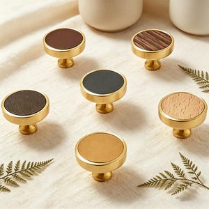 Può includere: Una collezione di pomelli per mobili in ottone con vari design superiori. I pomelli presentano una base con finitura dorata e top circolari in pelle marrone, pelle nera e motivi a venature del legno. I pomelli sono disposti su una superficie color crema.