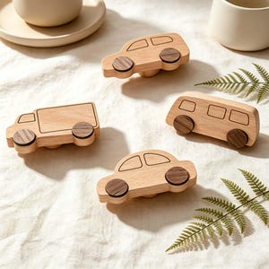 May include: Four wooden toy vehicles: a car, a van, a bus, and a truck. Each vehicle is made of light-colored wood with dark brown wooden wheels. The toys are arranged on a white surface, with a fern and a cup in the background.
