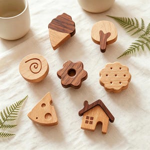 May include: A collection of wooden decorative knobs in various shapes, including an ice cream cone, tree, swirl, flower, cookie, cheese wedge, and house. The knobs are made of light and dark brown wood and are arranged on a white surface.