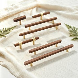 Puede incluir: Una colección de seis tiradores para armarios con barras de madera marrón oscuro y detalles en metal dorado cepillado. Los herrajes incluyen manijas horizontales de varias longitudes, sostenidas por soportes cilíndricos de color dorado.