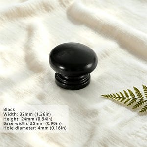 Puede incluir: Un pomo de mueble negro s&oacute;lido con una parte superior lisa y redondeada y una base escalonada. El pomo tiene un ancho de 32 mm, una altura de 24 mm, un ancho de base de 25 mm y un di&aacute;metro de orificio de 4 mm, seg&uacute;n el texto.