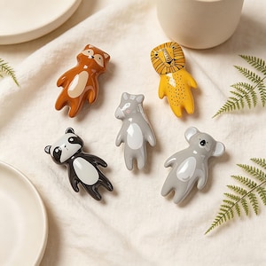 May include: A collection of ceramic animal-shaped chopstick rests. The set includes a fox, lion, wolf, panda, and koala, each with unique color schemes. The fox is brown, the lion is yellow, the wolf and koala are gray, and the panda is black and white.