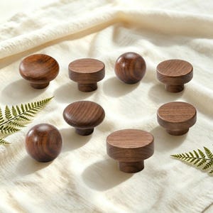 May include: A collection of eight dark brown wooden knobs with visible grain patterns. The set includes several cylindrical knobs with flat tops and two spherical knobs, all featuring a smooth, polished finish and a consistent natural wood texture.