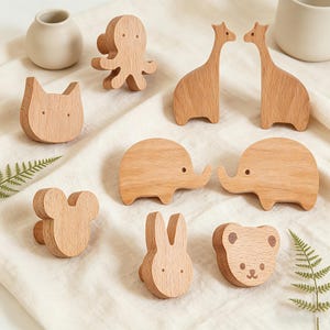 Può includere: Una collezione di figurine di animali in legno, tra cui un polpo, giraffe, elefanti, un gatto, un topo, un coniglio e un orso. Le figurine sono marroni chiare e hanno design semplici e arrotondati. Sono disposte su una superficie bianca.