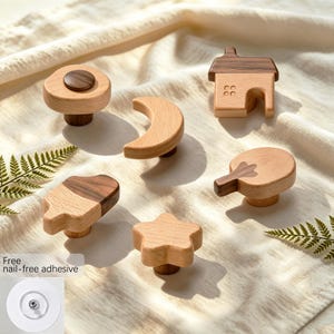 May include: A collection of six wooden cabinet knobs in various shapes including a house, a crescent moon, a star, a tree, and circular designs. The knobs feature light and dark wood tones and include a clear circular nail-free adhesive disc.