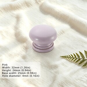 Puede incluir: Un pomo de mueble de color rosa claro con una parte superior redondeada y lisa y una base escalonada. El pomo tiene un ancho de 32 mm, una altura de 24 mm, un ancho de base de 25 mm y un di&aacute;metro de orificio de 4 mm, seg&uacute;n el texto.