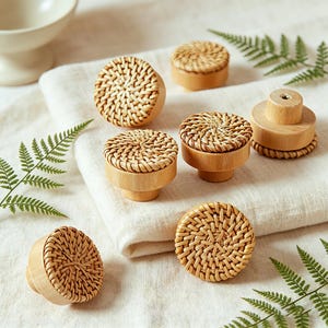 May include: A collection of wooden cabinet knobs with woven rattan tops. The knobs are in various shapes and sizes, with a natural wood finish. The knobs are arranged on a white cloth with green fern leaves.