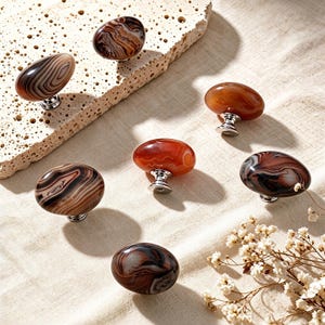 May include: A collection of seven oval-shaped cabinet knobs featuring polished, multi-colored agate stone tops with swirling patterns in shades of brown, black, orange, and cream, each mounted on a small, circular silver-toned metal base.