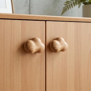 May include: Two light-colored wooden cabinet knobs with a rounded, cloud-like shape attached to a matching light wood cabinet door. The knobs feature a smooth, natural wood grain finish and are mounted side-by-side on the vertical cabinet panels.