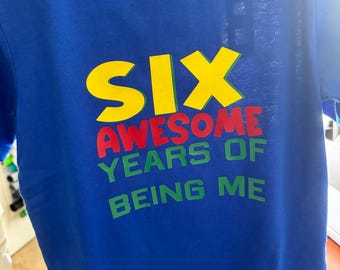 May include: Blue t-shirt with the text "SIX AWESOME YEARS OF BEING ME" in yellow, red, and green. The shirt has a crew neck and is made of a soft material.