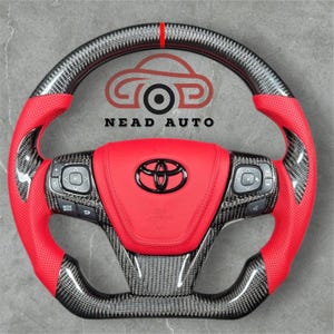 Op de afbeelding: Een aangepast autostuur met een rode lederen grip en een koolstofvezel afwerking. Het midden heeft een rood kussen met het Toyota-logo en de tekst "SRS AIRBAG". Het stuur heeft geïntegreerde bedieningselementen en de tekst "NEAD AUTO".