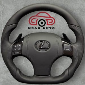Op de afbeelding: Zwart leren stuurwiel met het Lexus-logo in het midden. Het stuurwiel heeft geïntegreerde bedieningselementen en schakelflippers. De tekst "NEAD AUTO" en een autologo zijn bovenaan zichtbaar.