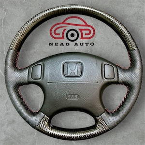 Può includere: Volante nero e in fibra di carbonio con cuciture rosse. Il volante presenta il logo Honda e il testo "NEAD AUTO". Il volante ha un'impugnatura in pelle traforata e un rivestimento in fibra di carbonio.