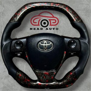 Puede incluir: Volante negro de Toyota con detalles en rojo y gris. El volante tiene un agarre de cuero perforado y controles integrados. El centro presenta el logotipo de Toyota. La imagen también incluye el texto "NEAD AUTO".