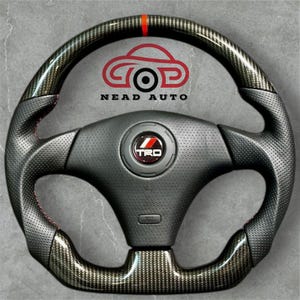 Peut inclure: Un volant noir et en fibre de carbone avec des coutures rouges et une bande rouge en haut. Le centre présente un logo TRD et le texte "NEAD AUTO".