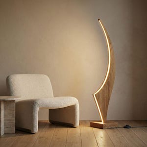 May include: A modern wooden floor lamp with a curved design and integrated LED lighting. The lamp has a natural wood finish and a warm, inviting glow. The lamp is placed next to a cream-colored chair.