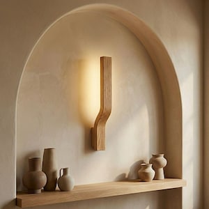 Puede incluir: Aplique de pared de madera con un diseño curvo, que emite un brillo cálido. El aplique está montado en una pared beige dentro de una alcoba arqueada. Una estantería de madera debajo exhibe varios jarrones de cerámica en tonos neutros.