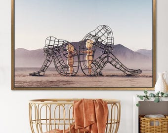 Può includere: Stampa artistica incorniciata con due figure a filo metallico in un paesaggio desertico. Le figure sono sedute, con la testa china, e racchiudono figure dorate più piccole. L'opera d'arte è ambientata su uno sfondo di montagne e un cielo azzurro pallido.