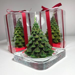 Könnte beinhalten: Grüne Weihnachtsbaumkerzen mit goldenen Glitzerakzenten, präsentiert in transparenten Geschenkboxen mit rotem Band. Die Kerzen stehen auf einem transparenten Glas-Kerzenhalter. Festliche Weihnachtsdekoration.