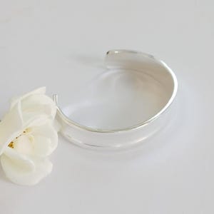May include: A polished silver-toned cuff bracelet with a smooth, wide band and an open back design, positioned next to a single white rose with delicate petals and a green stem, showcasing a clean, reflective metallic surface and floral detail.
