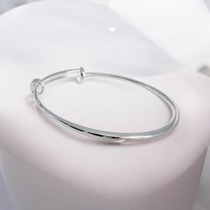 May include: A polished silver-tone bangle bracelet featuring a smooth, rounded band and a bypass-style overlapping closure mechanism. The metal has a reflective, metallic finish that catches the light, creating a clean and simple circular silhouette.