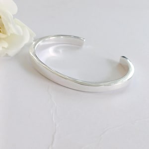 May include: A polished silver cuff bracelet with a smooth, reflective surface and an open-ended design. The bracelet features a simple, curved band shape and is positioned next to a white flower petal, highlighting its metallic finish.
