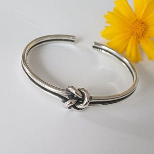 May include: A polished silver cuff bracelet featuring a central knot design. The bracelet has an open back and a smooth, reflective metallic finish. A bright yellow flower is positioned near the open end of the silver band.