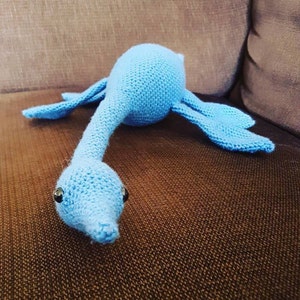Könnte beinhalten: Ein blaues, gehäkeltes Plüschtier in Form eines langhalsigen Dinosauriers mit großen Augen. Das Spielzeug liegt auf einem braunen Sofa.