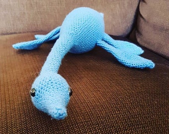 May include: A blue crocheted plush toy shaped like a long-necked dinosaur with large eyes. The toy is lying on a brown couch.