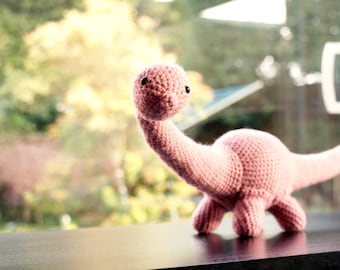 May include: A pink crocheted dinosaur toy with black eyes. The dinosaur is standing on a dark surface.