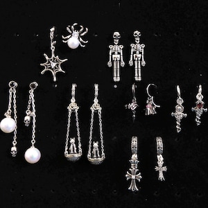 Puede incluir: Colección de pendientes plateados con diversos diseños: arañas, telarañas, esqueletos, calaveras, perlas y cruces. Se exhiben sobre un fondo negro, mostrando una temática gótica o de Halloween.
