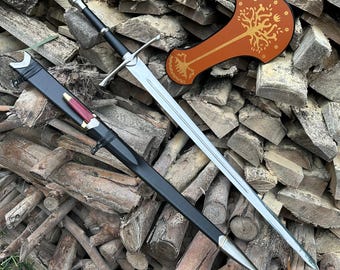Puede incluir: Una espada de estilo medieval con una hoja plateada y un mango negro, apoyada sobre una pila de madera. Una vaina negra con detalles plateados y un pequeño mango rojo y plateado está junto a la espada. También hay un escudo marrón con un diseño de árbol dorado.