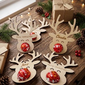Puede incluir: Cuatro adornos de madera clara con forma de reno, cada uno con un recorte redondo que sostiene una bola de chocolate envuelta en papel rojo como nariz. Los adornos están grabados con nombres o frases como Frohe Weihnachten, Claudia, Jens y Merry Christmas.