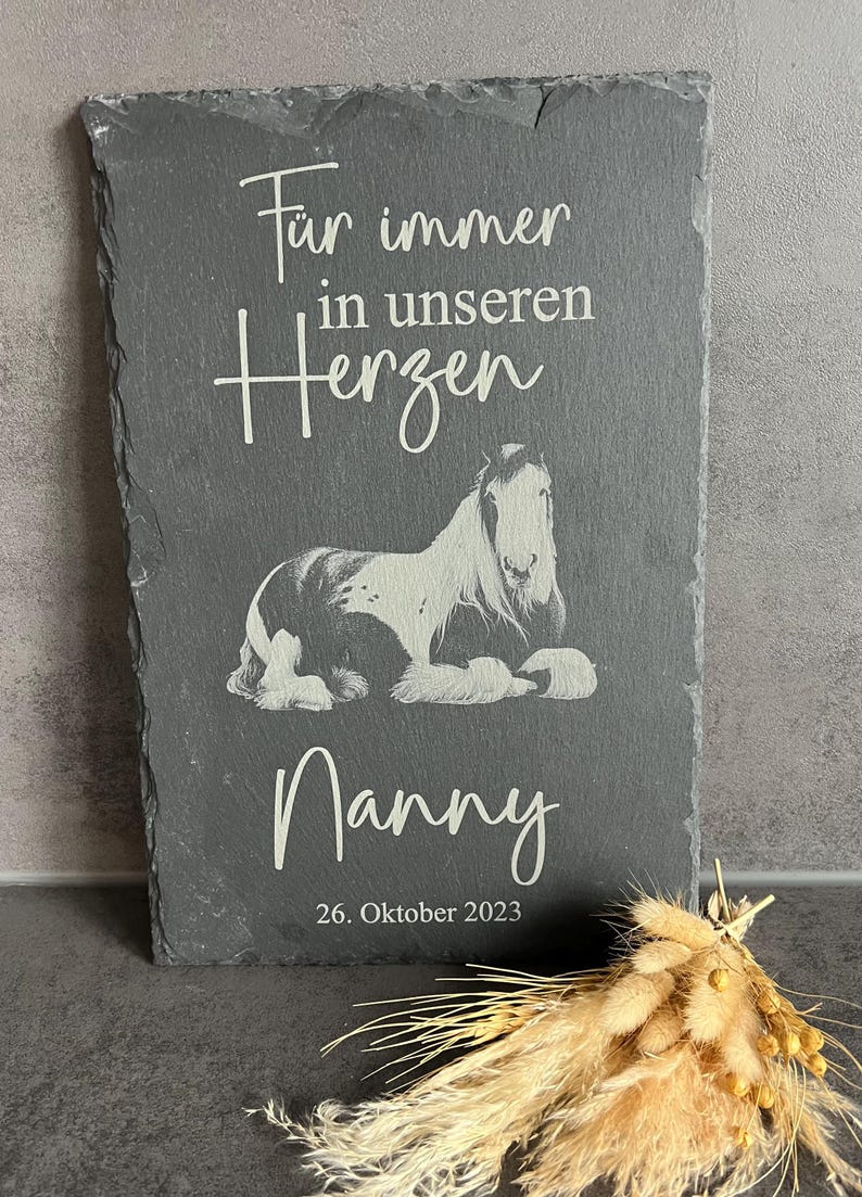 Puede incluir: Una placa rectangular de pizarra gris oscuro con la imagen grabada de un caballo tumbado. El texto dice "F&uuml;r immer in unseren Herzen", "Nanny" y "26. Oktober 2023". La placa tiene un borde de piedra natural rugoso y una superficie plana.