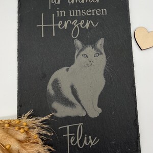 Puede incluir: Una placa rectangular de pizarra negra con la imagen grabada de un gato en el centro. El texto "F&uuml;r immer in unseren Herzen" est&aacute; grabado sobre el gato y el nombre "Felix" debajo. A la derecha hay un peque&ntilde;o coraz&oacute;n de madera.