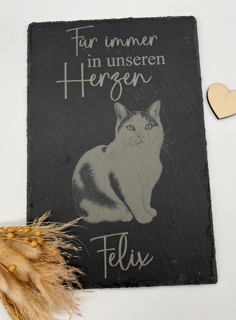 Puede incluir: Una placa rectangular de pizarra negra con la imagen grabada de un gato en el centro. El texto "F&uuml;r immer in unseren Herzen" est&aacute; grabado sobre el gato y el nombre "Felix" debajo. A la derecha hay un peque&ntilde;o coraz&oacute;n de madera.