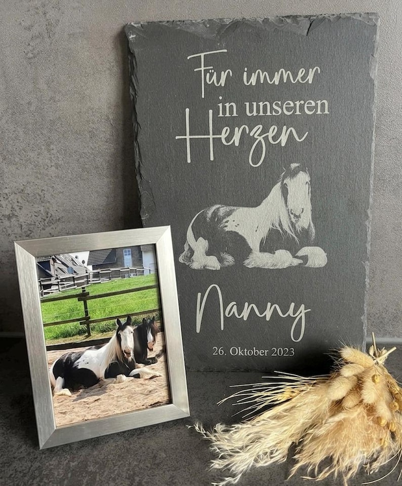 Puede incluir: Una placa rectangular de pizarra gris oscuro grabada con el texto "F&uuml;r immer in unseren Herzen", "Nanny" y "26. Oktober 2023" junto a una ilustraci&oacute;n de un caballo, acompa&ntilde;ada de un marco plateado con una foto de dos caballos y hierba seca.