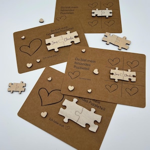 Puede incluir: Una colección de piezas de rompecabezas y tarjetas de madera personalizadas. Las tarjetas son marrones con ilustraciones de corazones y el texto "Du bist mein fehlendes Puzzleteil" y "Ich liebe dich". Las piezas del rompecabezas están grabadas con nombres.