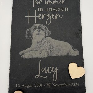 Puede incluir: Una placa rectangular de pizarra negra con la imagen grabada de un perro tumbado. El texto dice "F&uuml;r immer in unseren Herzen", "Lucy" y "12. August 2008 - 28. November 2023". Se incluyen dos peque&ntilde;os corazones de madera clara.