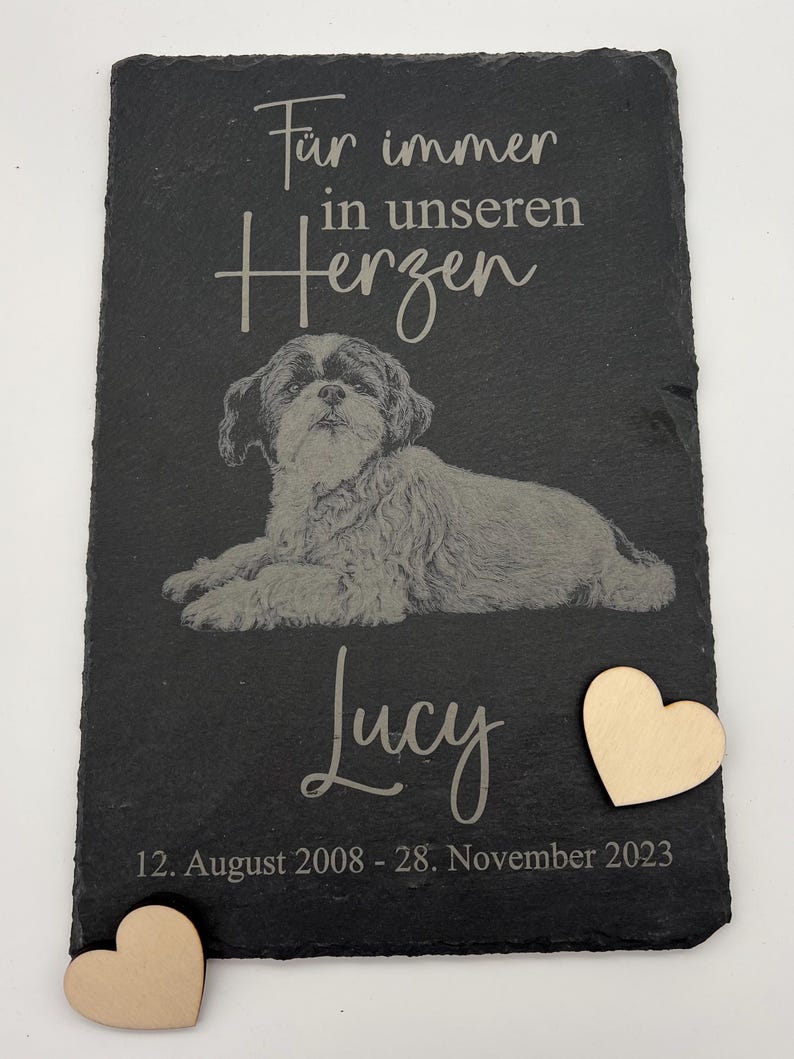 Puede incluir: Una placa rectangular de pizarra negra con la imagen grabada de un perro tumbado. El texto dice "F&uuml;r immer in unseren Herzen", "Lucy" y "12. August 2008 - 28. November 2023". Se incluyen dos peque&ntilde;os corazones de madera clara.