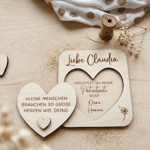 Könnte beinhalten: Eine helle Holzplatte mit herzförmigem Ausschnitt und ein separates herzförmiges Holzstück. Der Text lautet: Liebe Claudia, MÖCHTEST DU MEINE Patentante SEIN? Deine Hanna, und KLEINE MENSCHEN BRAUCHEN SO GROBE HERZEN WIE DEINS.